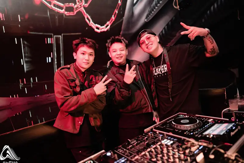 Ba DJ tạo dáng sau bàn mixer của ALPHA Nha Trang, dây xích lớn treo phía trên bốt DJ