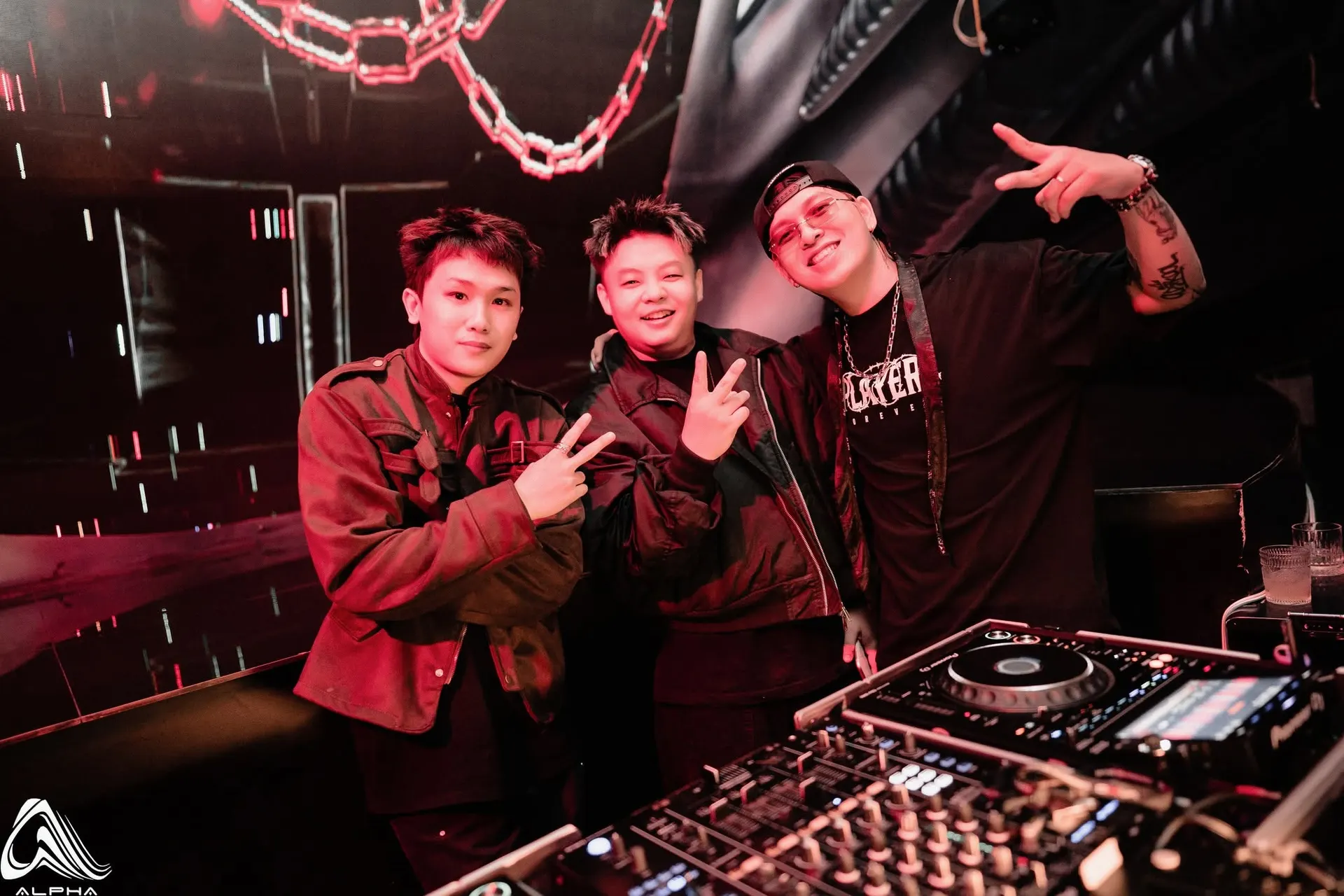 Ba DJ tạo dáng sau bàn mixer của ALPHA Nha Trang, dây xích lớn treo phía trên bốt DJ